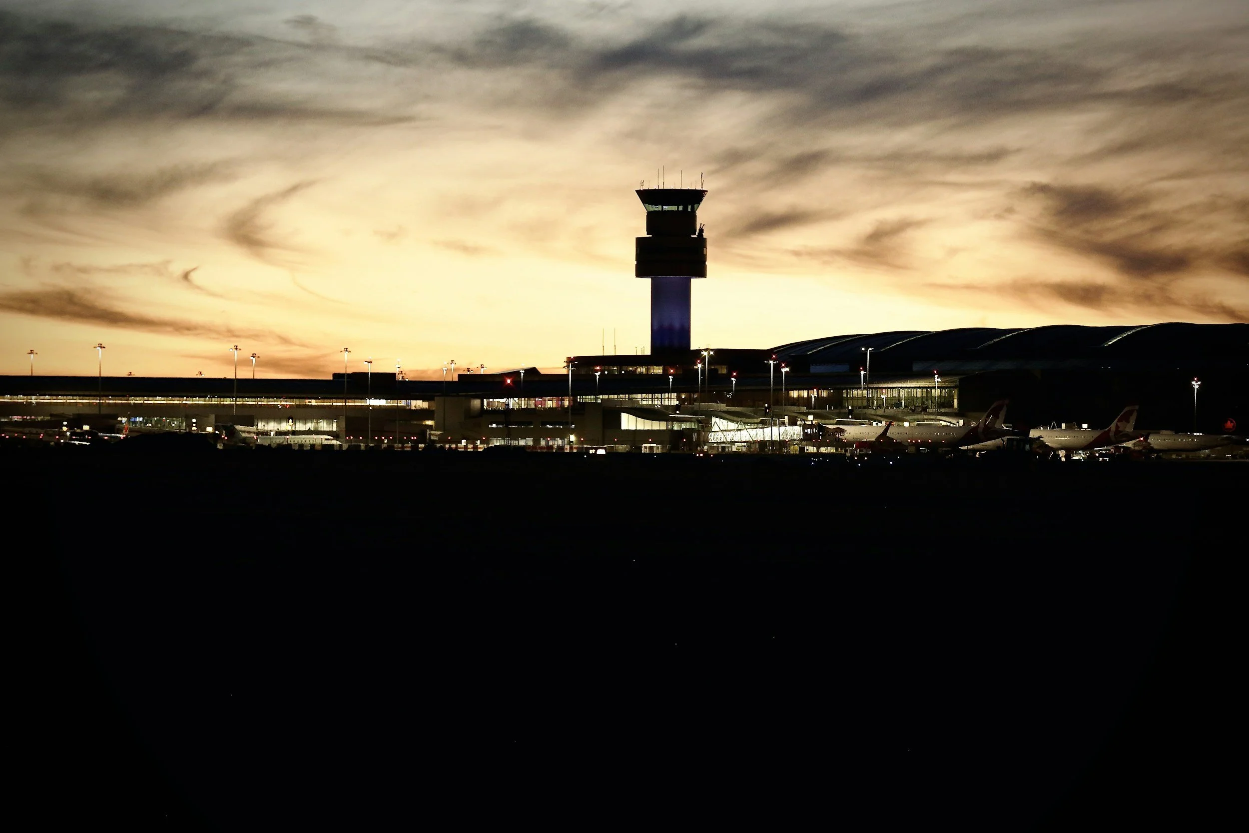 “Night view of airport with parked airplanes and control tower, under cloudy sky and warm sunset glow — ready for late arrivals and early pickups.”