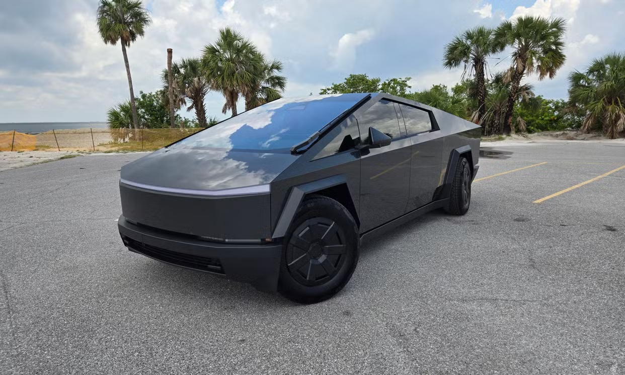 A black futuristic electric vehicle parked on an empty parking lot near a beach, with palm trees and cloudy sky in the background.