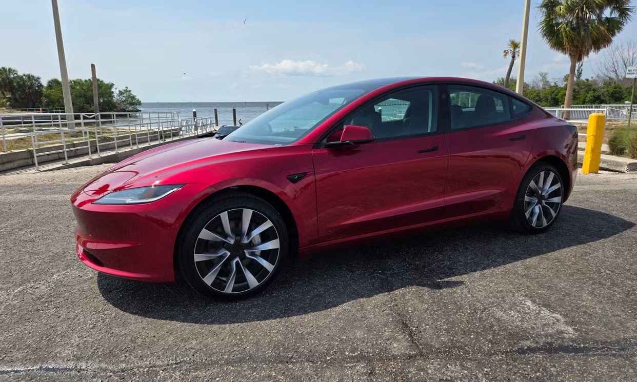 Red Tesla Model 3 parked by the water, with palm trees and metal railings in a tropical Florida setting.