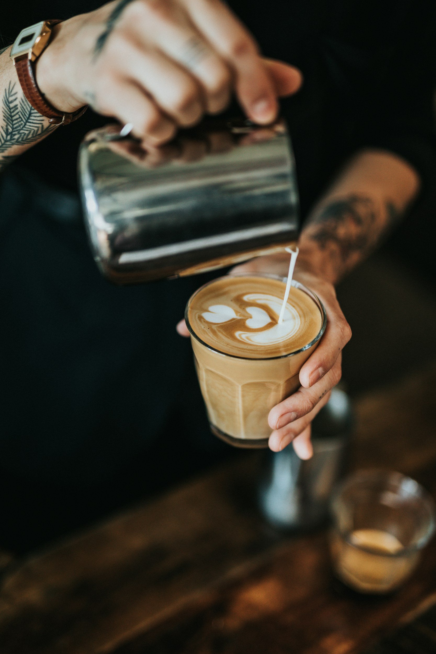 Person pouring milk into a latte with latte art in a glass cup
