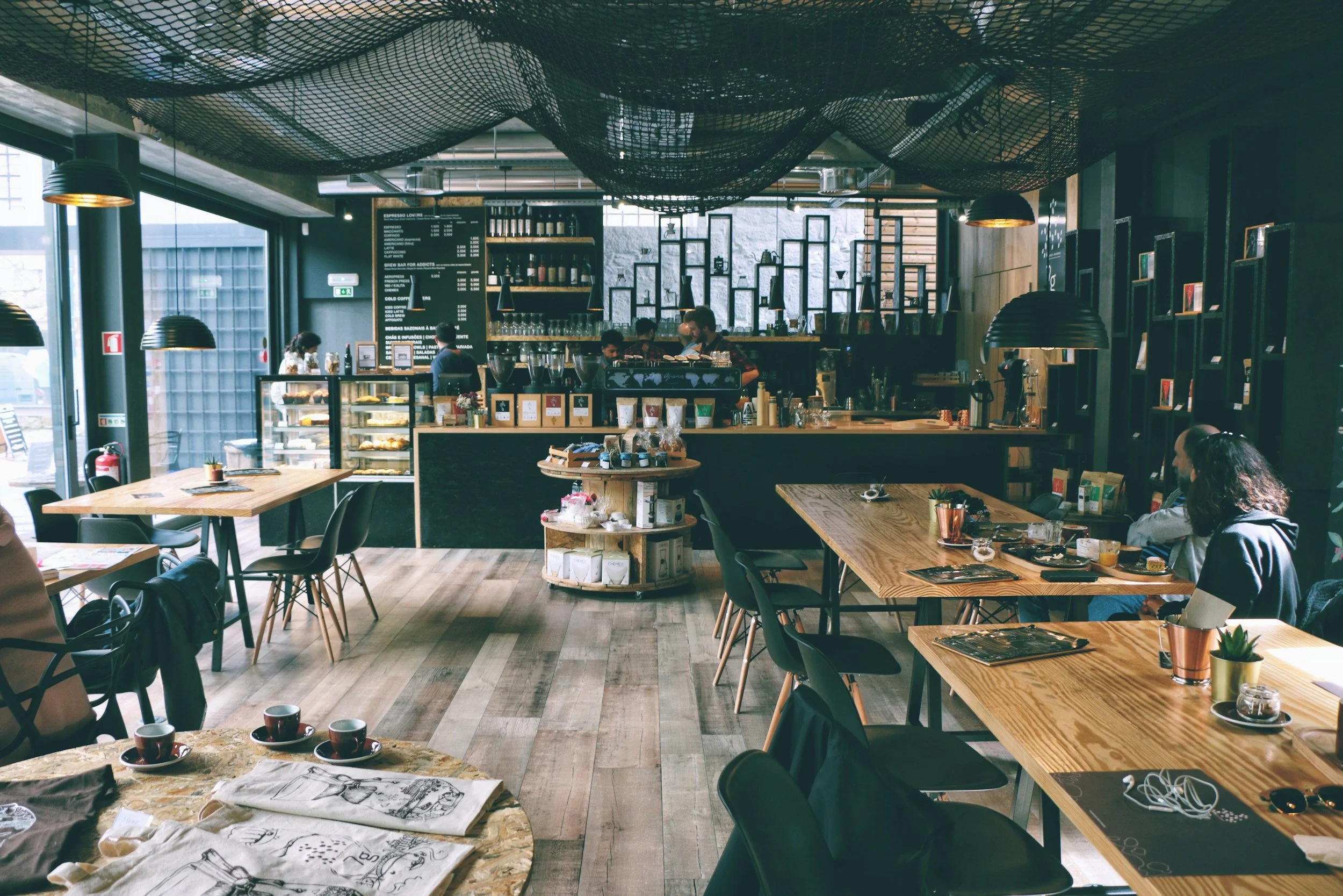 Interior of a modern coffee shop with wooden tables, black chairs, hanging pendant lights, a counter with pastries and a menu, and customers seated and ordering.