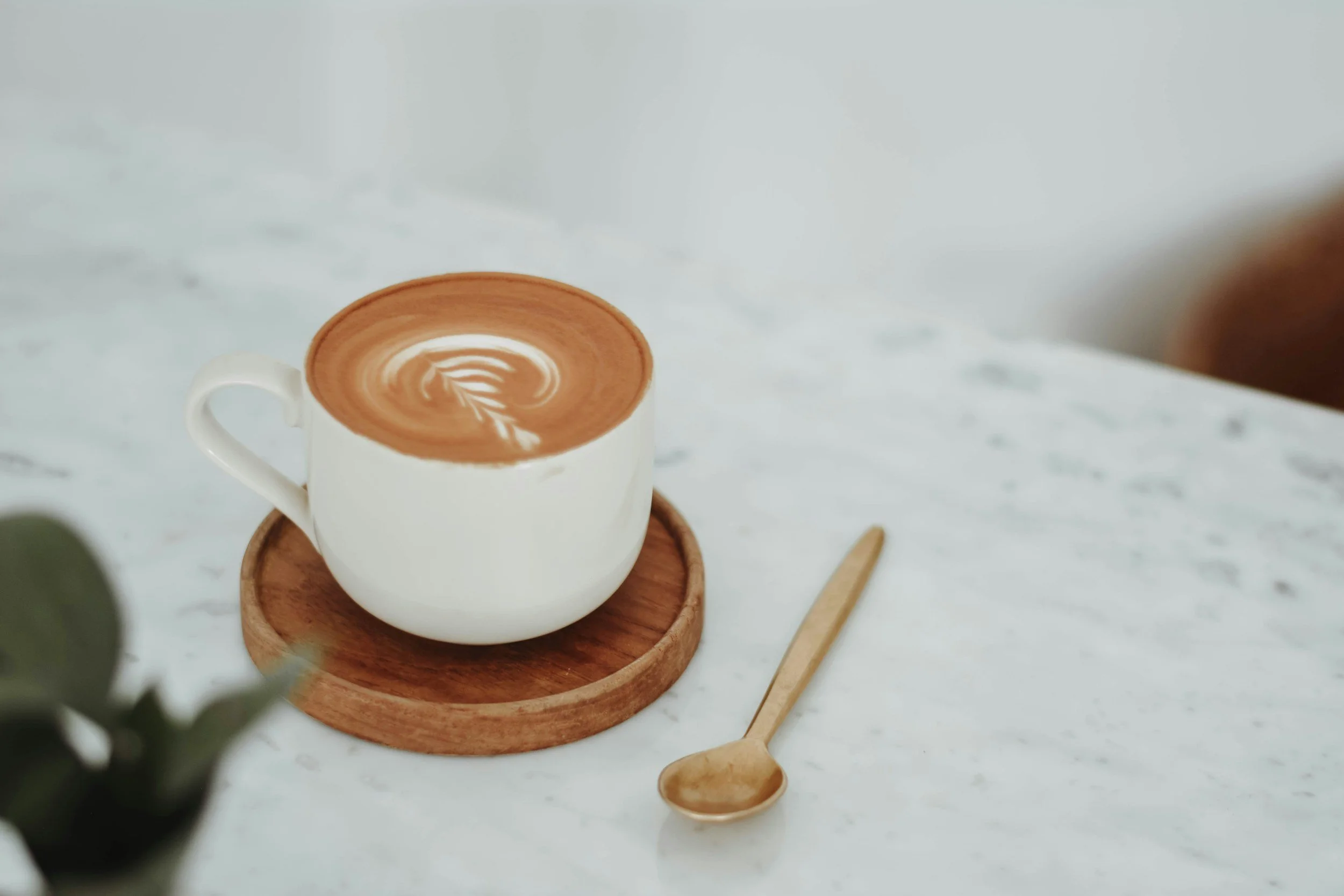 A cup of latte with foam art on top placed on a wooden coaster on a white marble table, with a small brass spoon nearby.