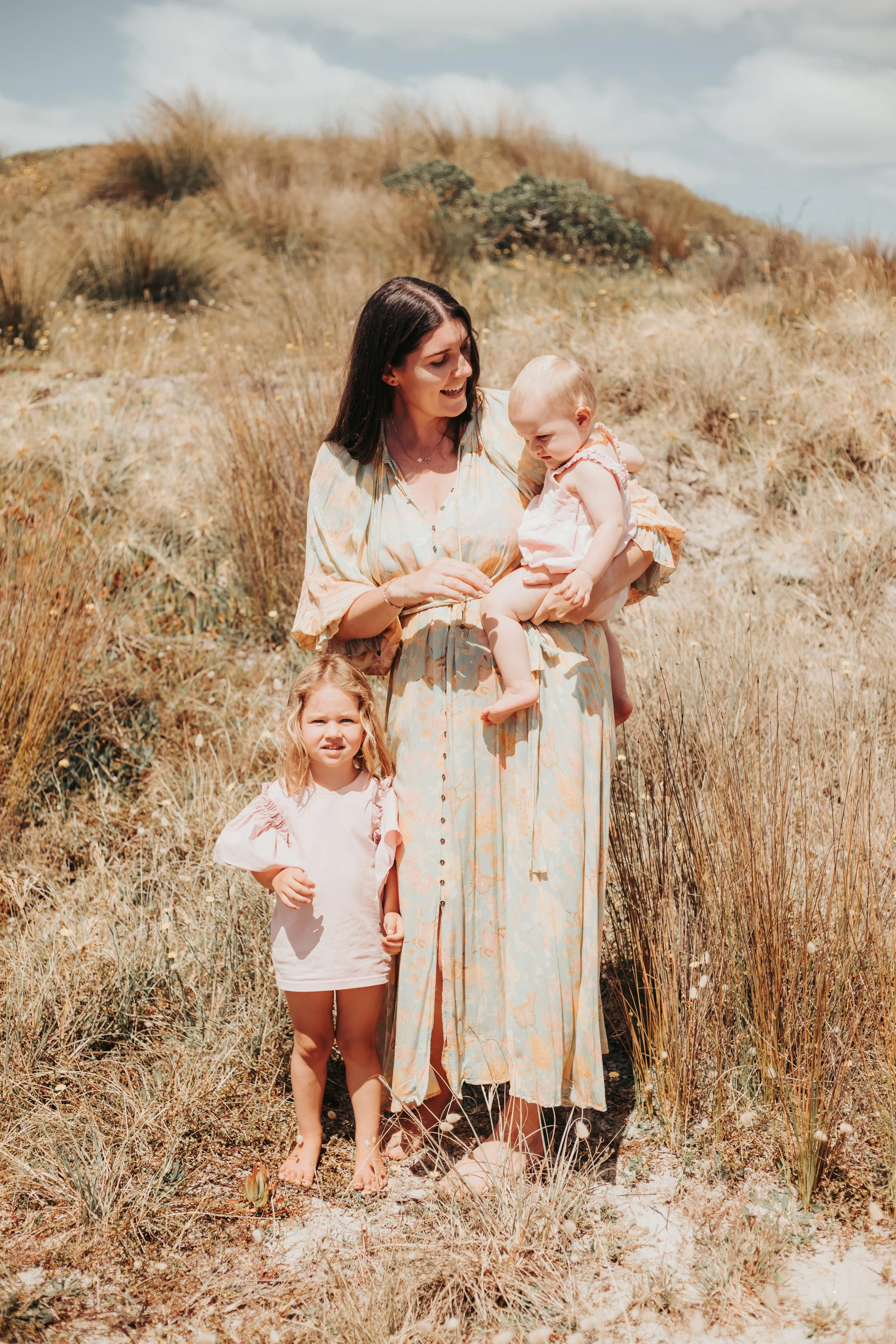 A woman holding a baby girl in a light-colored dress, with a young girl standing nearby, outdoors in a grassy, sandy area with dry vegetation.