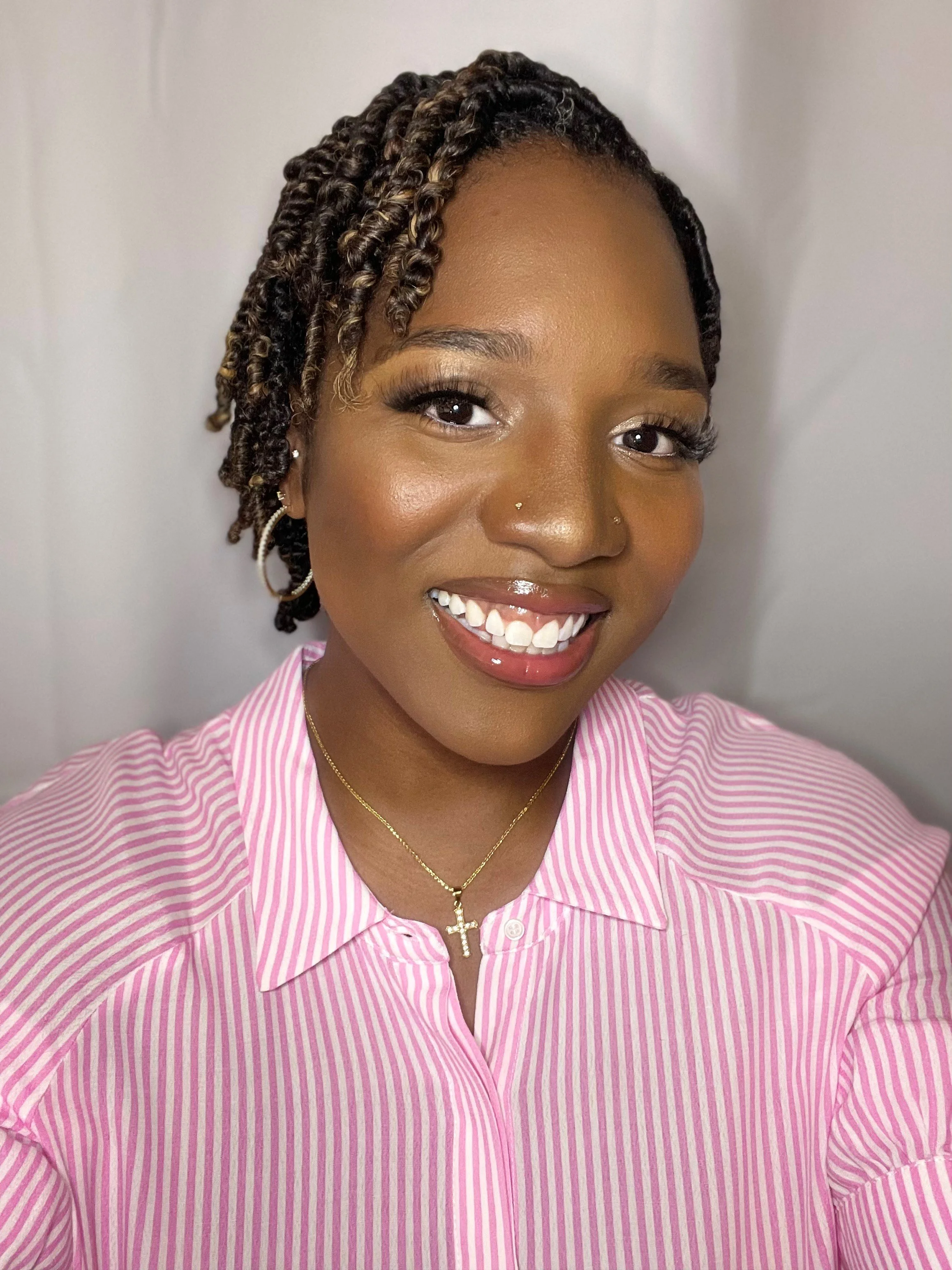 A woman with dark skin, curly hair styled in twists, smiling, wearing a pink and white striped shirt, gold necklace with a cross pendant, and hoop earrings.