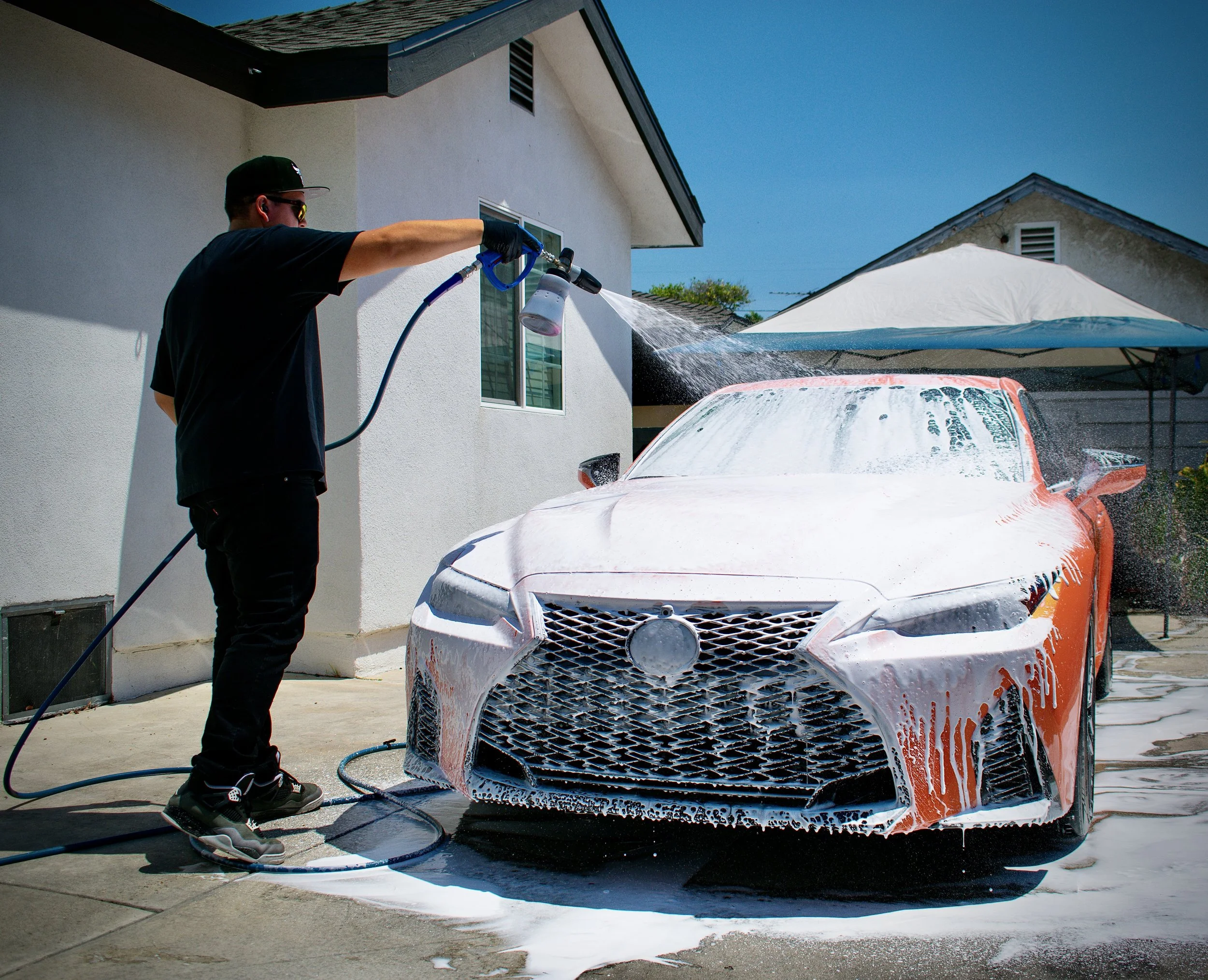 Person rinsing an orange car with a hose and soap outside a house.