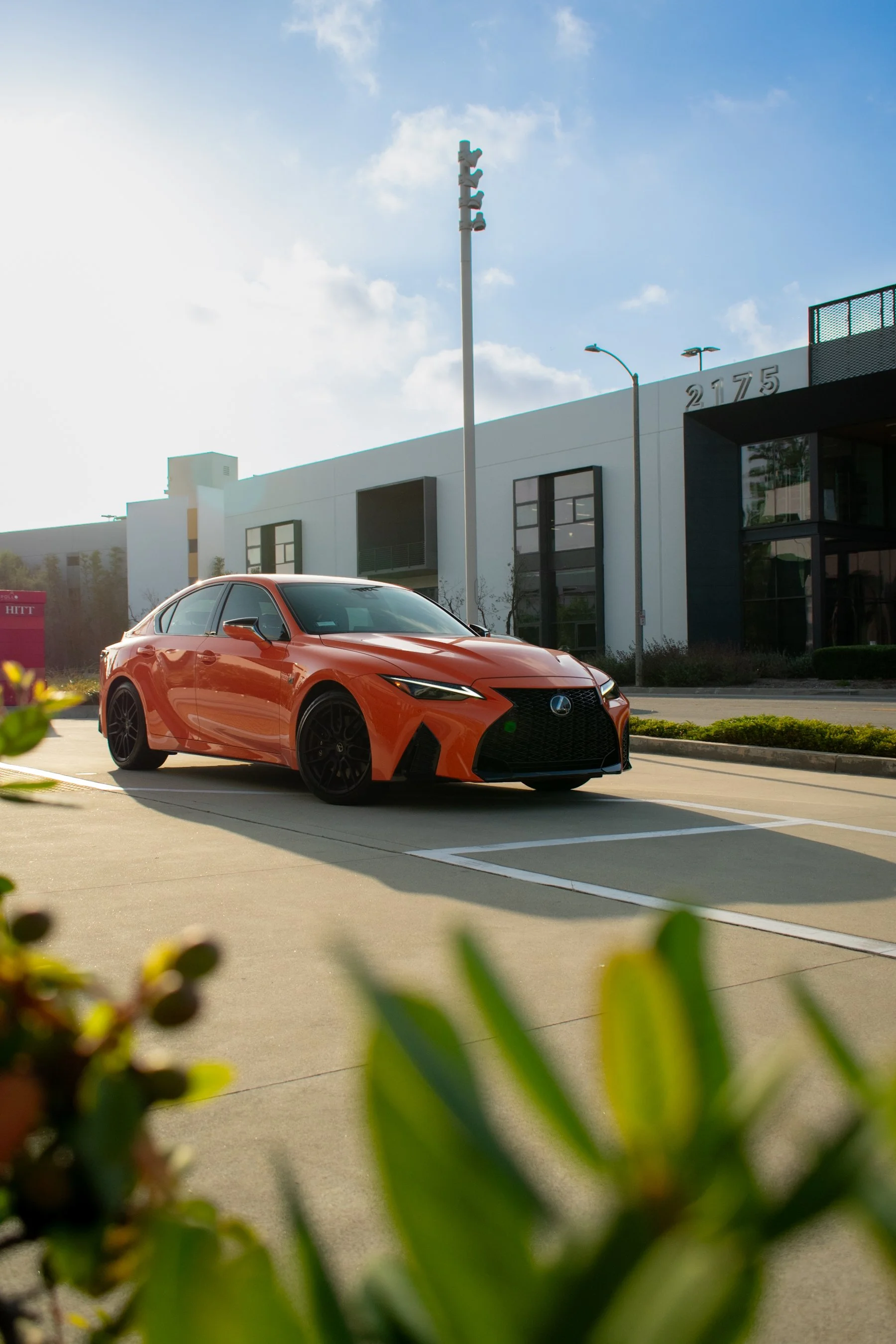 An orange Lexus sedan parked in an outdoor parking lot next to a modern building with large windows, under a partly cloudy sky.