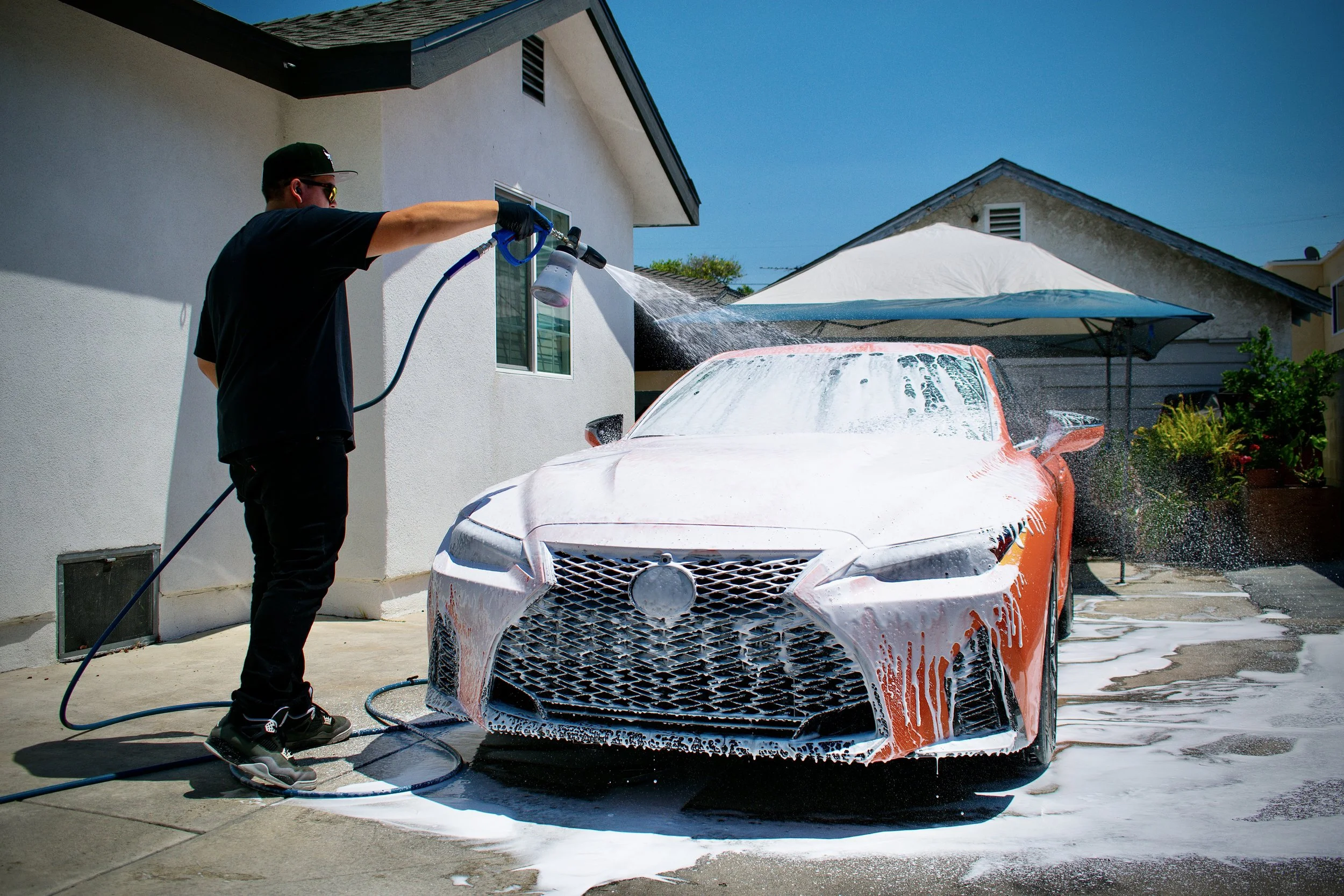 A man washing a bright orange sports car with soap and water outside a house under a patio umbrella on a sunny day.