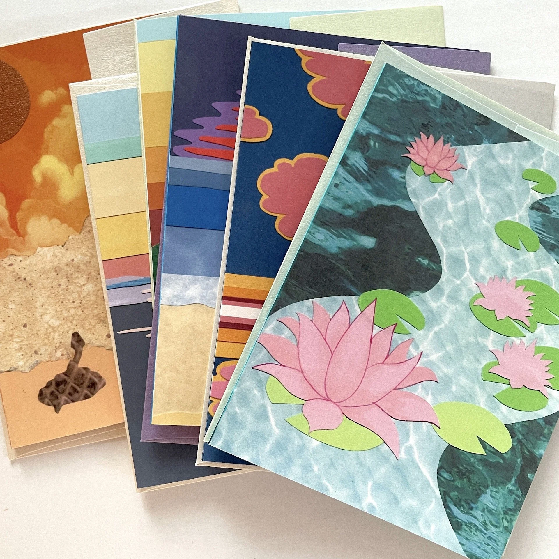 A selection of colorful greeting cards featuring various designs, including water lilies with pink flowers and green leaves, abstract clouds with a sunset, a snake in the desert, a seascape, and vibrant geometric patterns, with shiny envelopes.