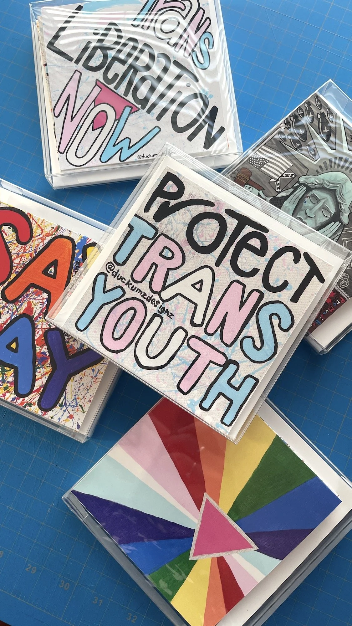 Colorful handmade protest signs with messages like 'HANDS LIBERATION NOW' and 'PROTECT TRANS YOUTH', displayed in clear plastic sleeves, on a blue cutting mat.