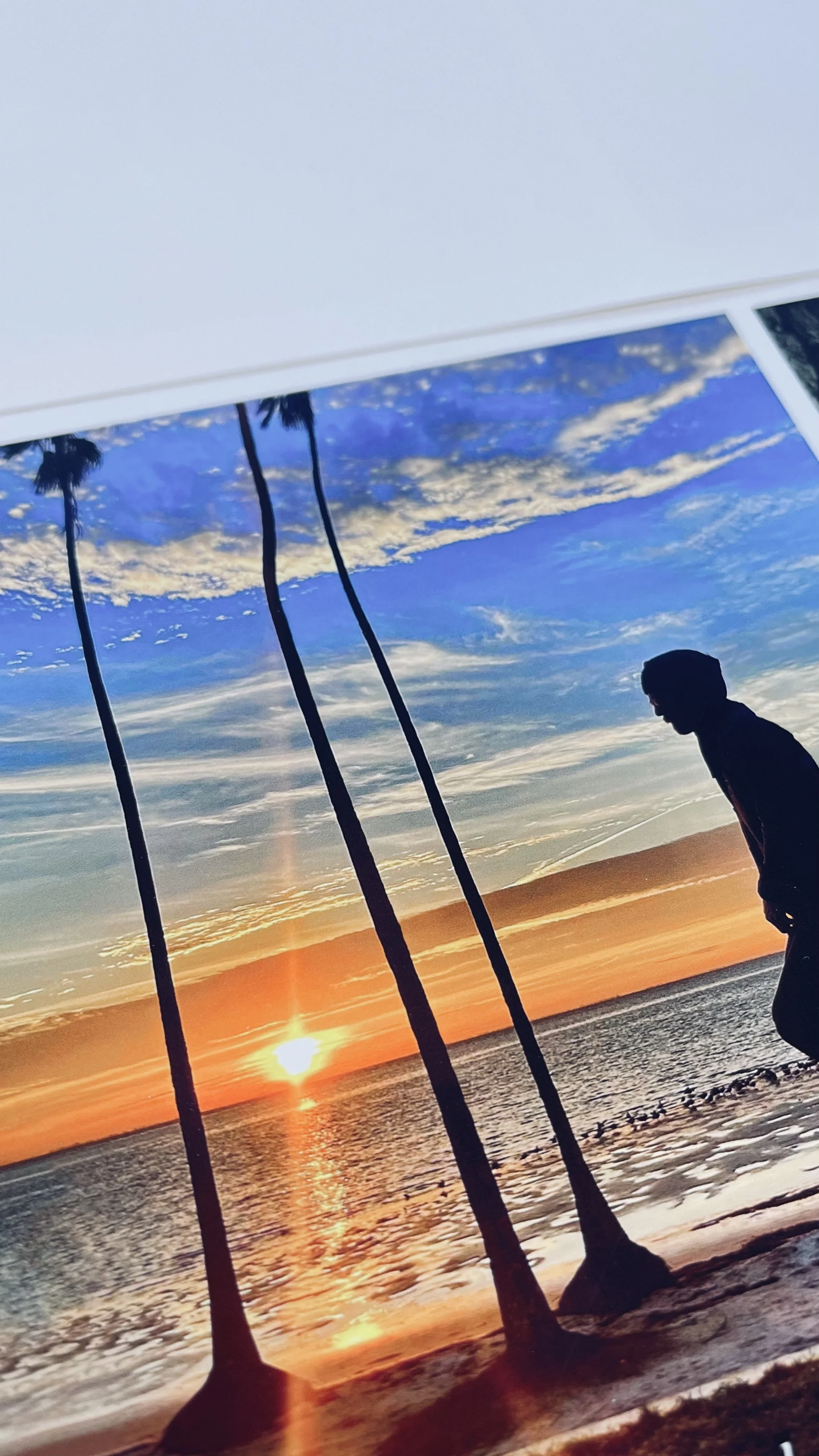 Close-up of a fine art print on bright white matte paper. Red River Polar Matte. The artwork is a vivid photograph depicting a silhouette of a person riding a unicycle on a beach at sunset with three tall palm trees and rippling waves.