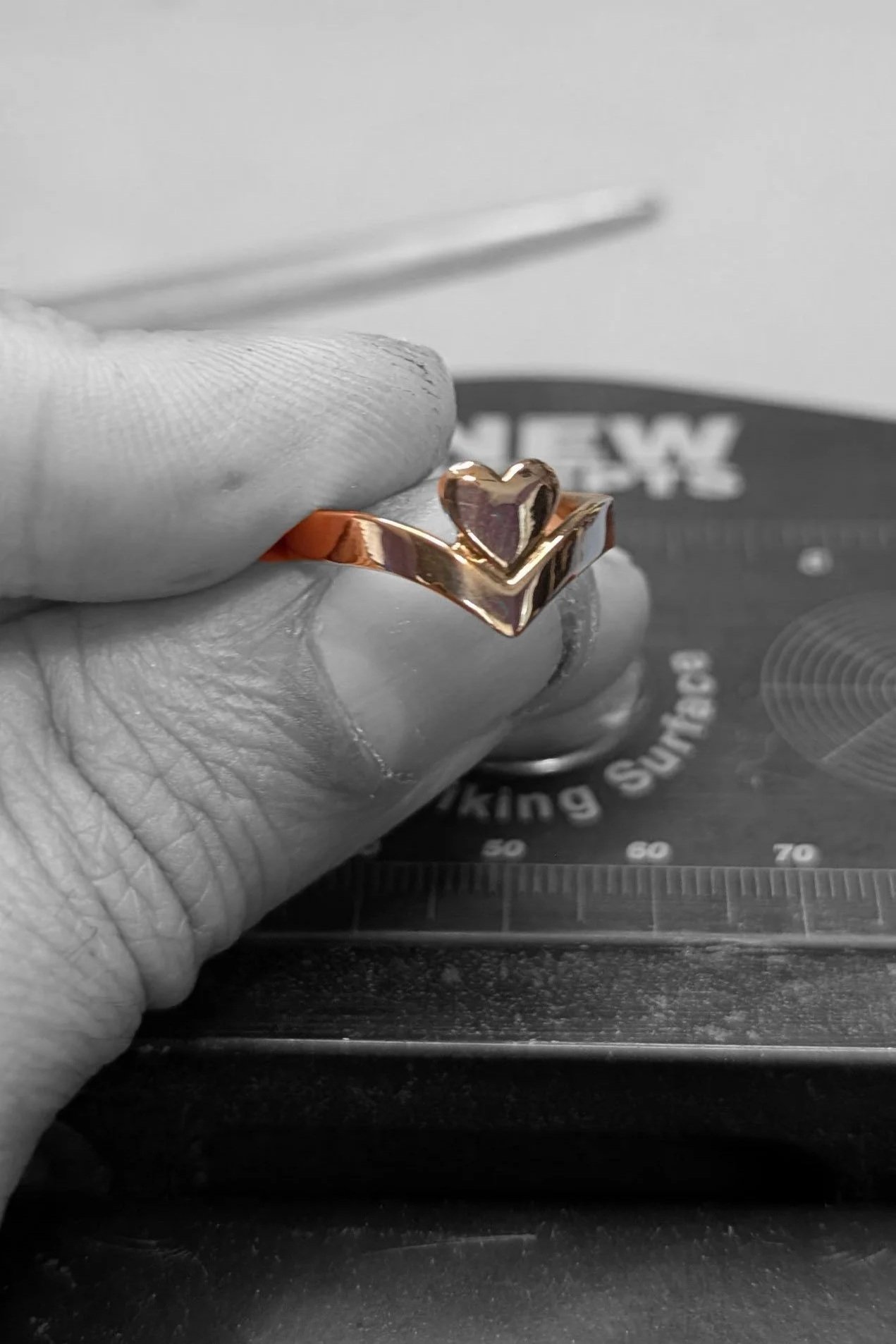 Close-up of a person's hand holding a gold ring with a heart-shaped centerpiece, in front of a jewelry craftsmanship background.