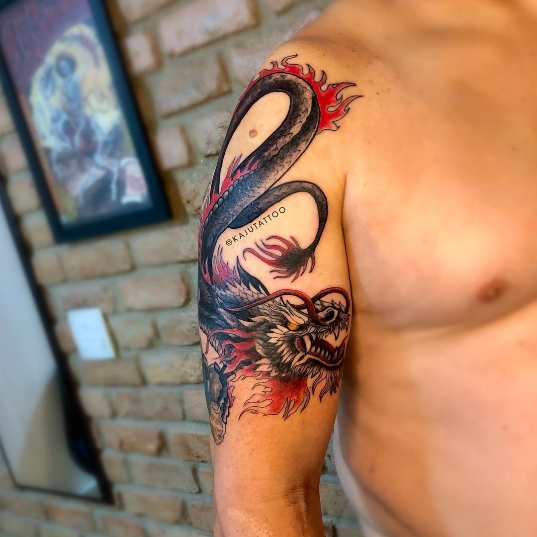 Tatuagem de um dragão com cabeça de leão na parte superior do braço, com detalhes em preto, vermelho e cinza.