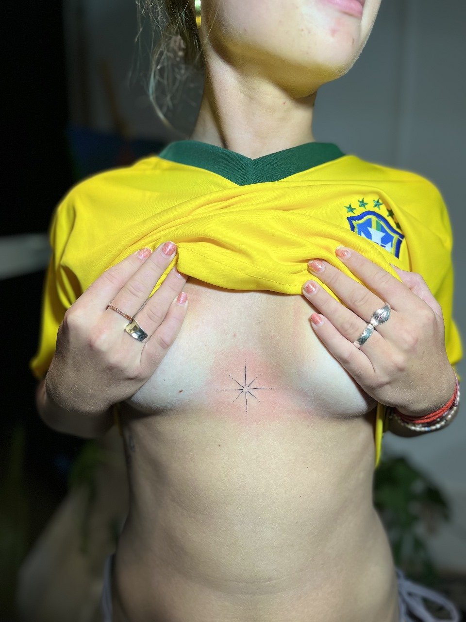 Pessoa com camiseta amarela da seleção brasileira de futebol levantando a blusa, expondo o seio e uma tatuagem pequena no centro do peito.