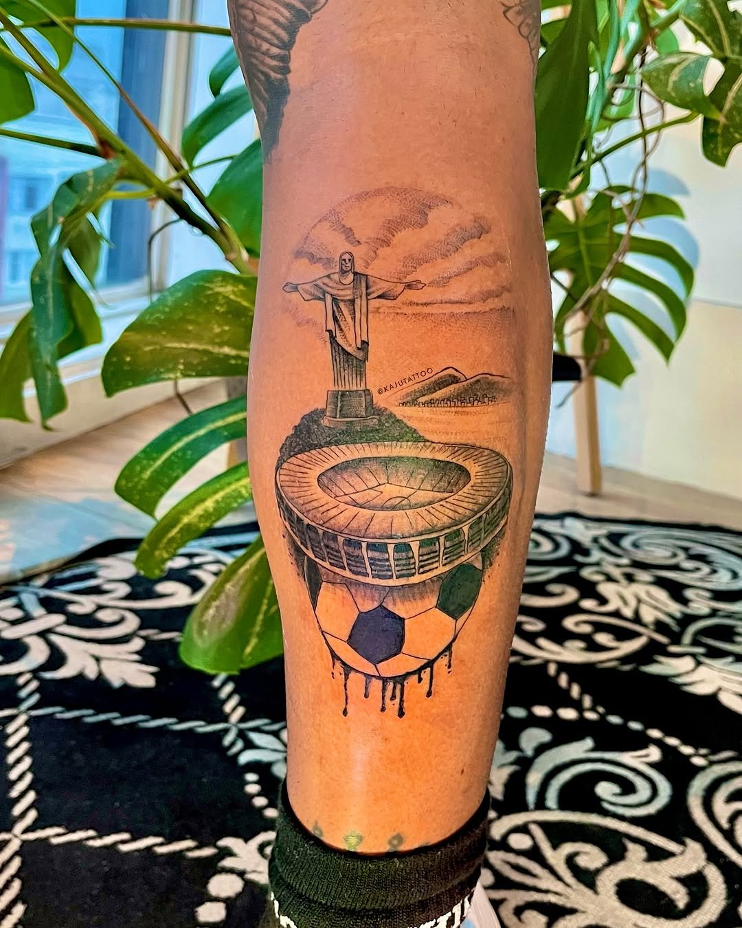 Tattoo de uma estátua parecida com o Cristo Redentor, sobre um estádio de futebol, com um futebol na base, tudo em preto e cinza.