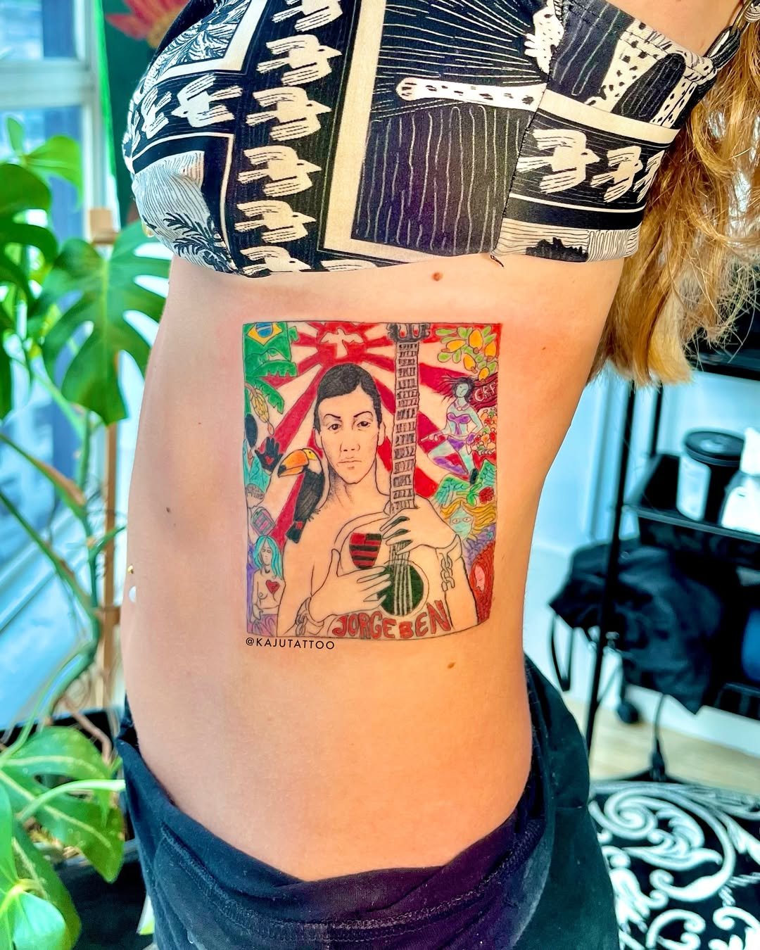 Tattoo colorido de uma personagem com cabelo curto e mãos segurando uma guitarra, com o nome 'Jorge Ben' na parte inferior, cercada por elementos da natureza e animais.