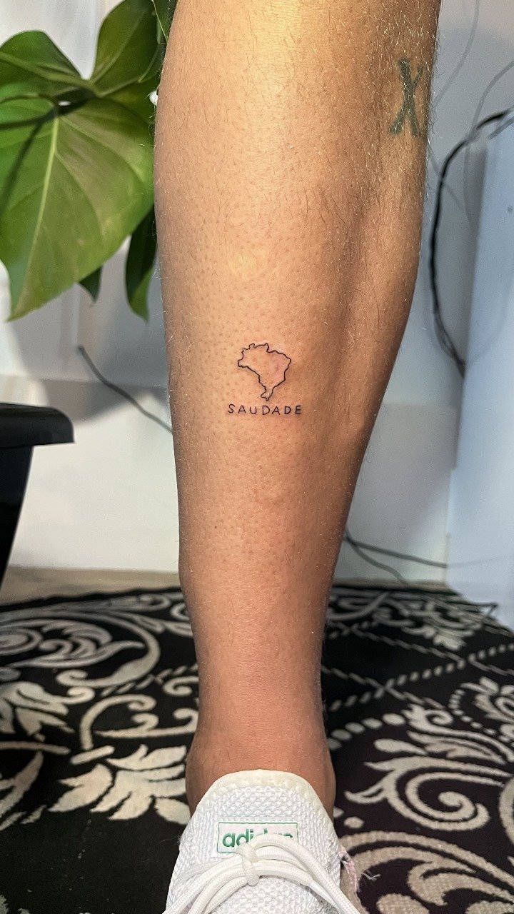 Tatuagem de mapa do Brasil com a palavra 'Saudade' na perna de uma pessoa