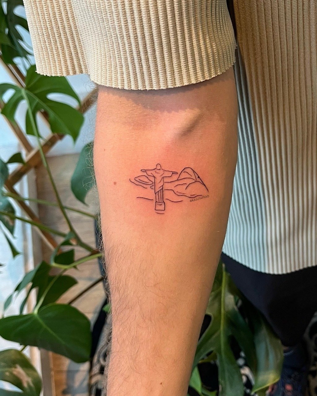 Tatuagem de uma pessoa em estilo minimalista de um homem de braços abertos em frente a uma paisagem montanhosa.