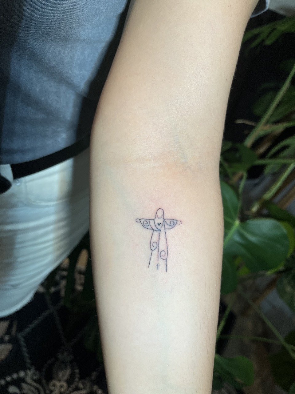 Tatuagem de uma figura delicada em forma de cruz, com detalhes curvilíneos, localizada no antebraço de uma pessoa.