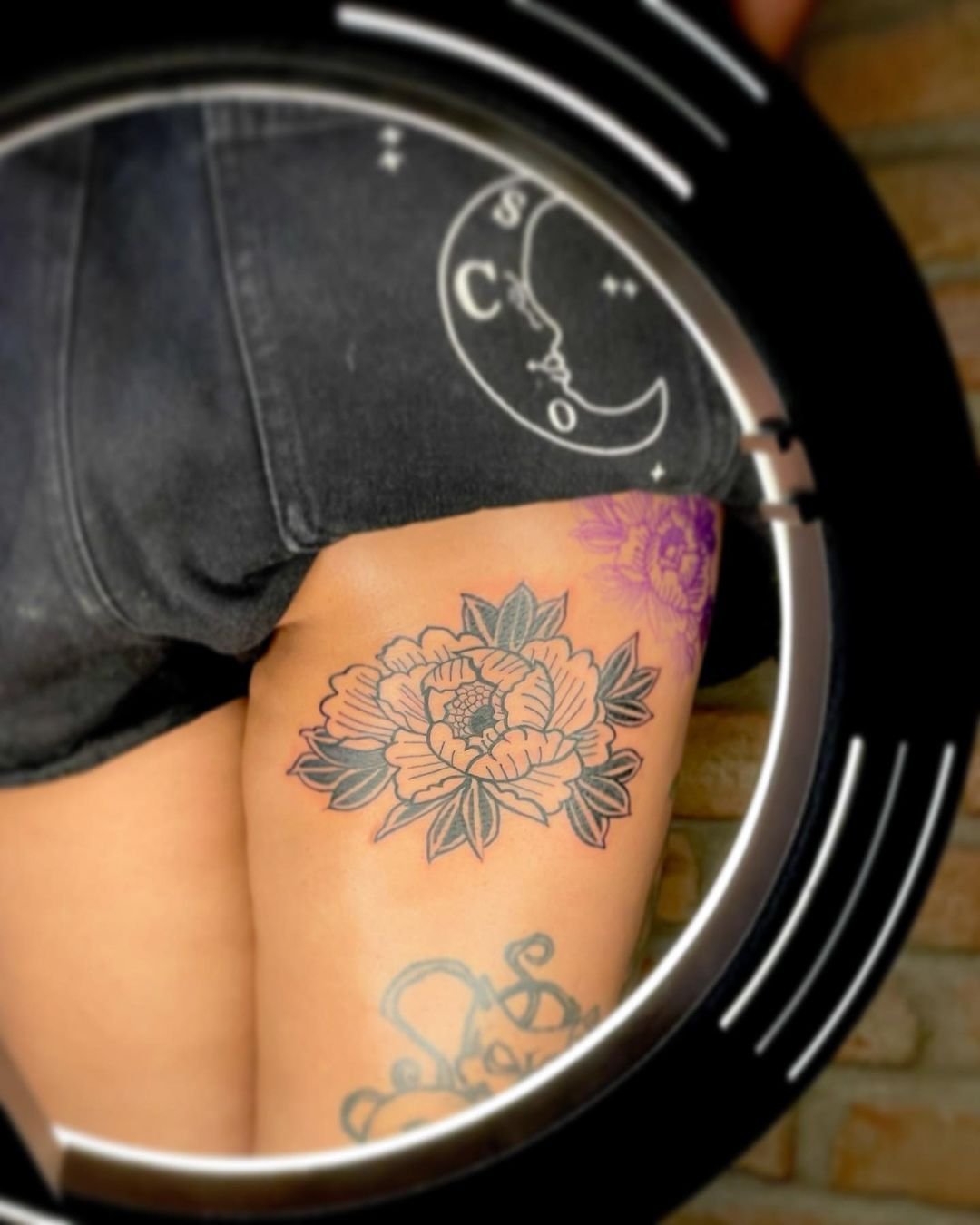 Tattoo de uma flor de peônia com folhas na coxa de uma pessoa, vista com um espelho redondo.