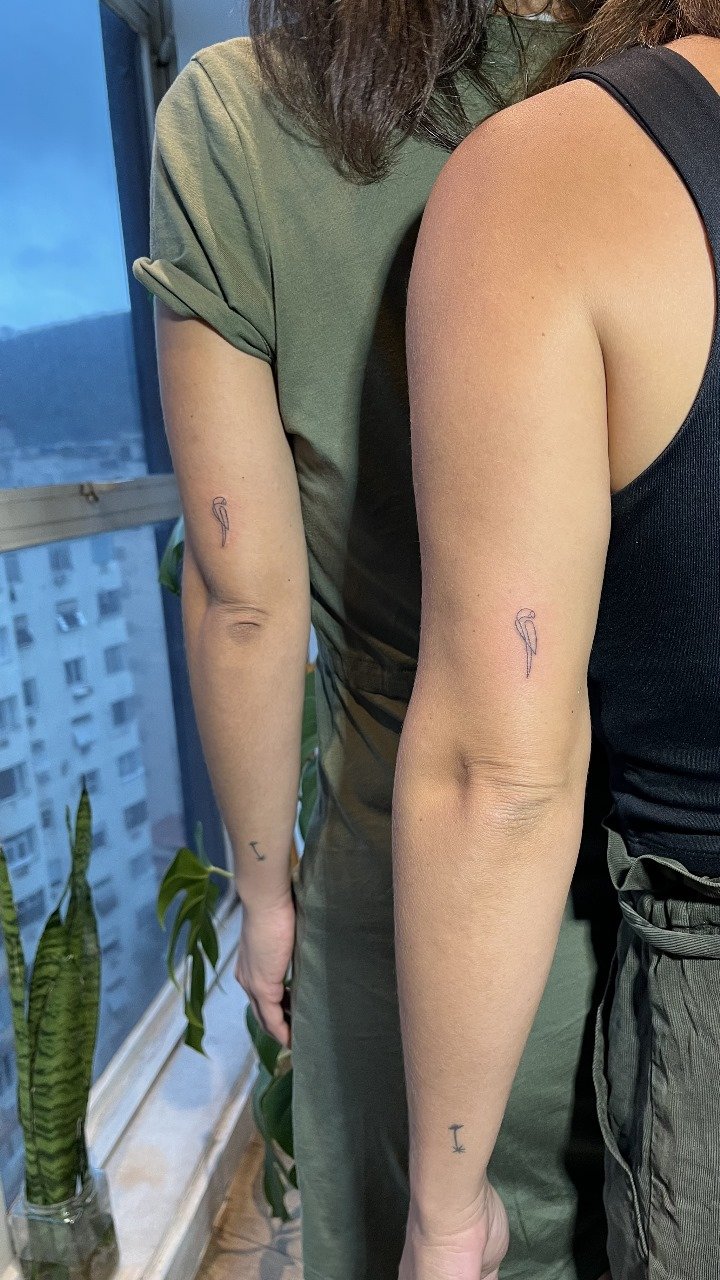 Duas pessoas com tatuagens de flamingo no braço, uma ao lado da outra, em ambiente interno próximo a uma janela.