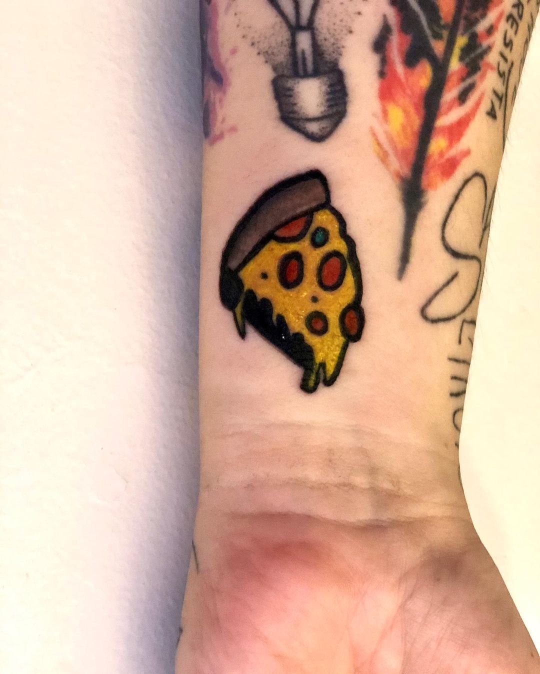 Tatuação de uma fatia de pizza de pepperoni com uma parte de queijo e massa visíveis, em cores vibrantes, localizada no antebraço de uma pessoa.