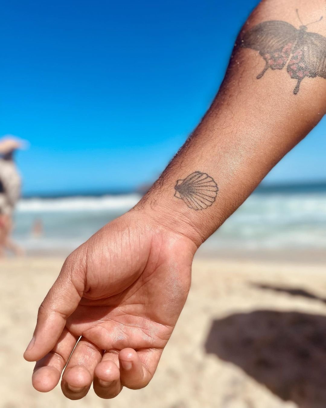 Mão de pessoa com tatuagens, uma delas de uma concha e outra de uma abelha, segurando a mão de outra pessoa na praia, com o mar e o céu azul ao fundo.