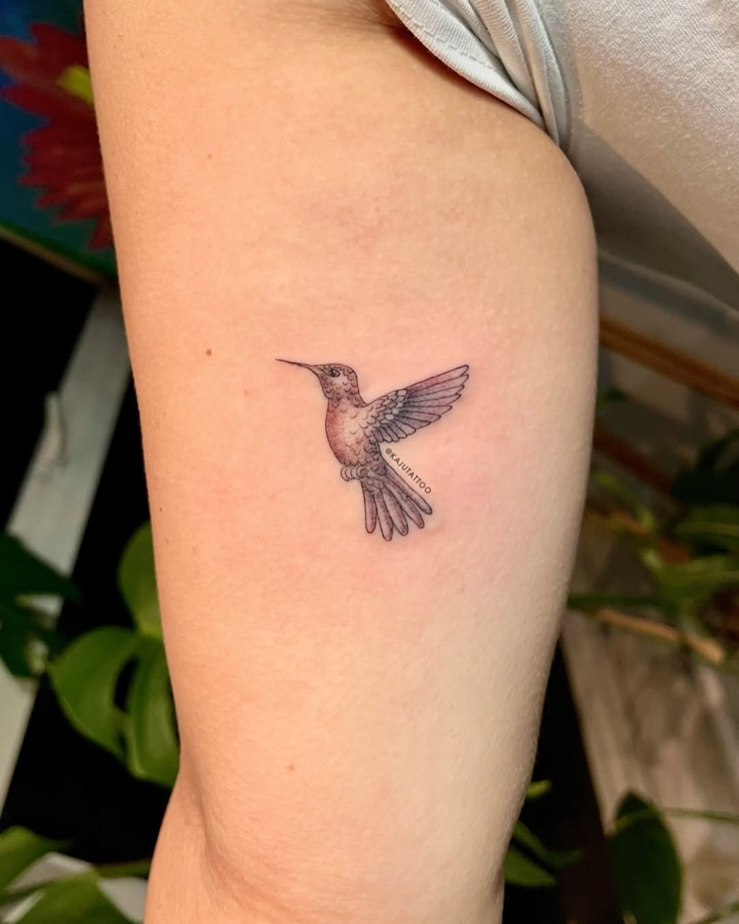Tattoo de um beija-flor minimalista na parte superior do braço, com detalhes em linhas finas e sombreamento suave.