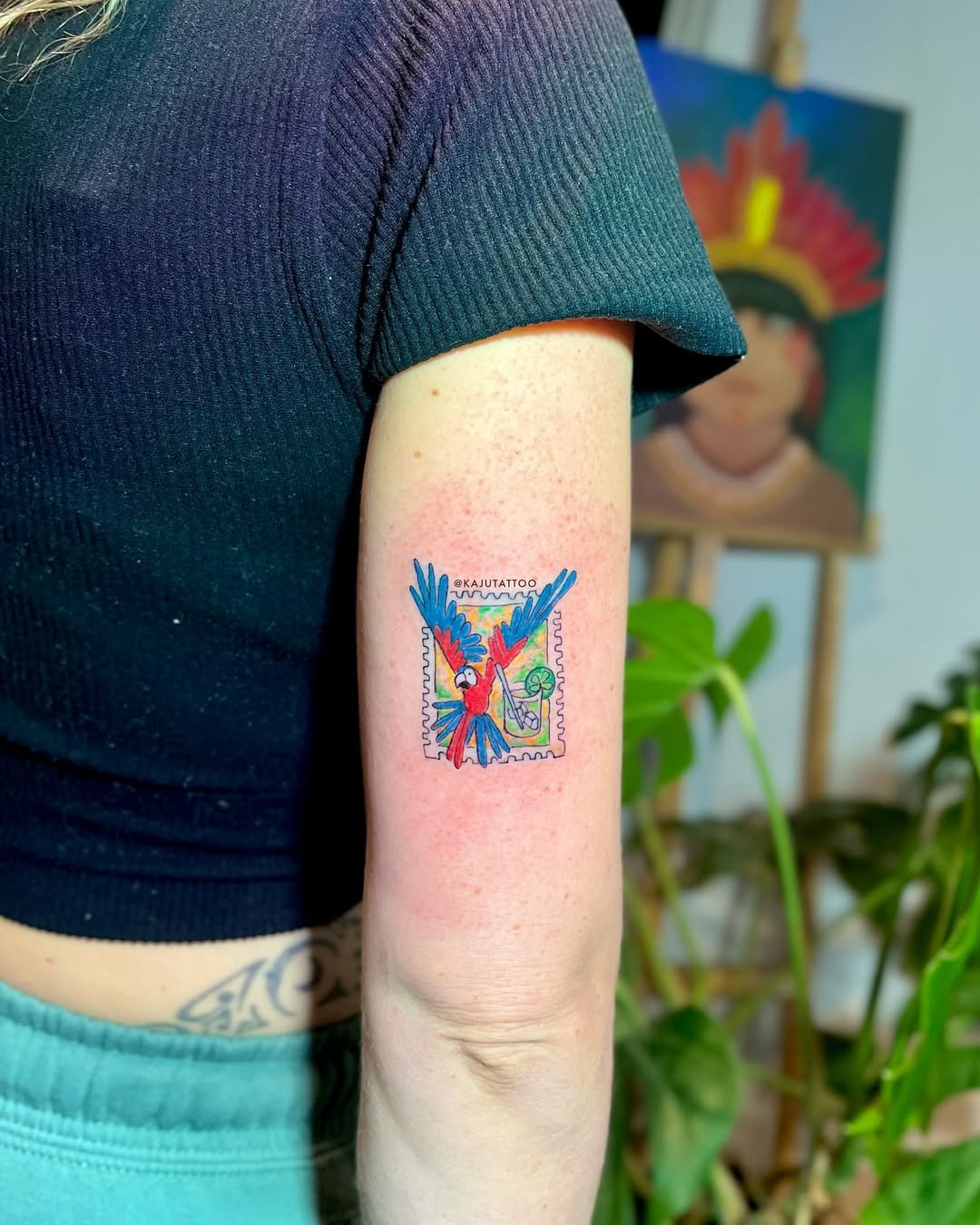 Tatuagem de um papagaio colorido em um braço, com detalhes de uma unha postiça com cores vibrantes e fundo de plantas e uma pintura ao fundo.