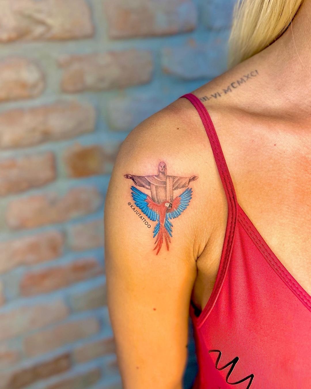 Tatuagem de uma cruz com um tucano colorido na frente do braço de uma mulher que usa uma blusa vermelha. No fundo, uma parede de tijolos.