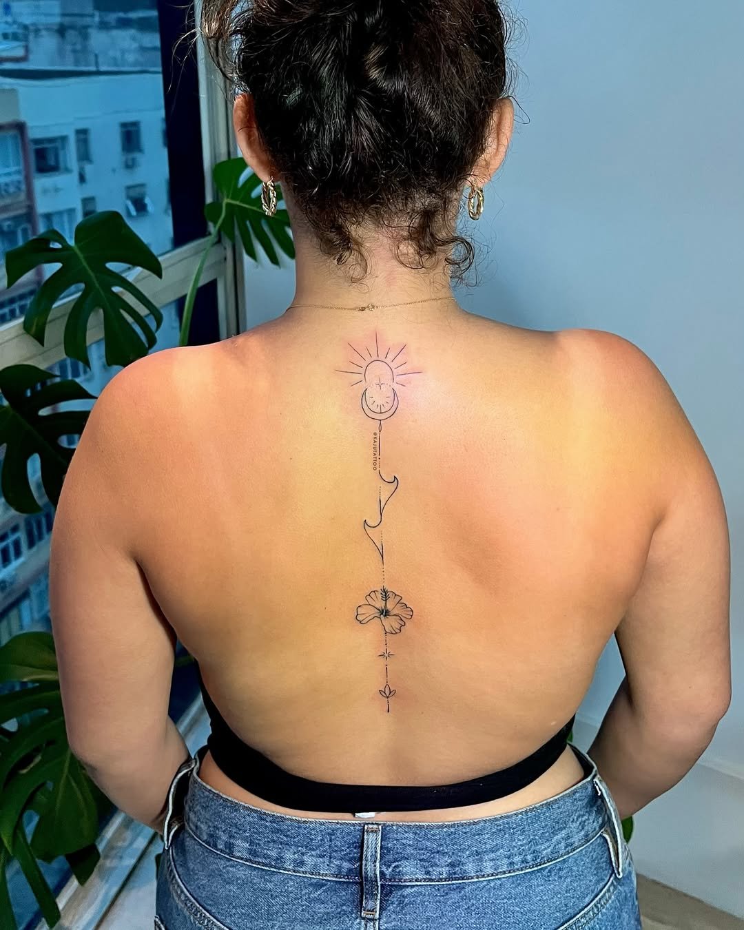Tatuagem de uma borboleta, uma linha pontilhada com um fio, uma lua crescente, linhas de raios de sol e linhas decorativas na parte superior das costas de uma mulher de cabelo castanho escuro e pele clara, usando brincos de argola e roupas jeans.