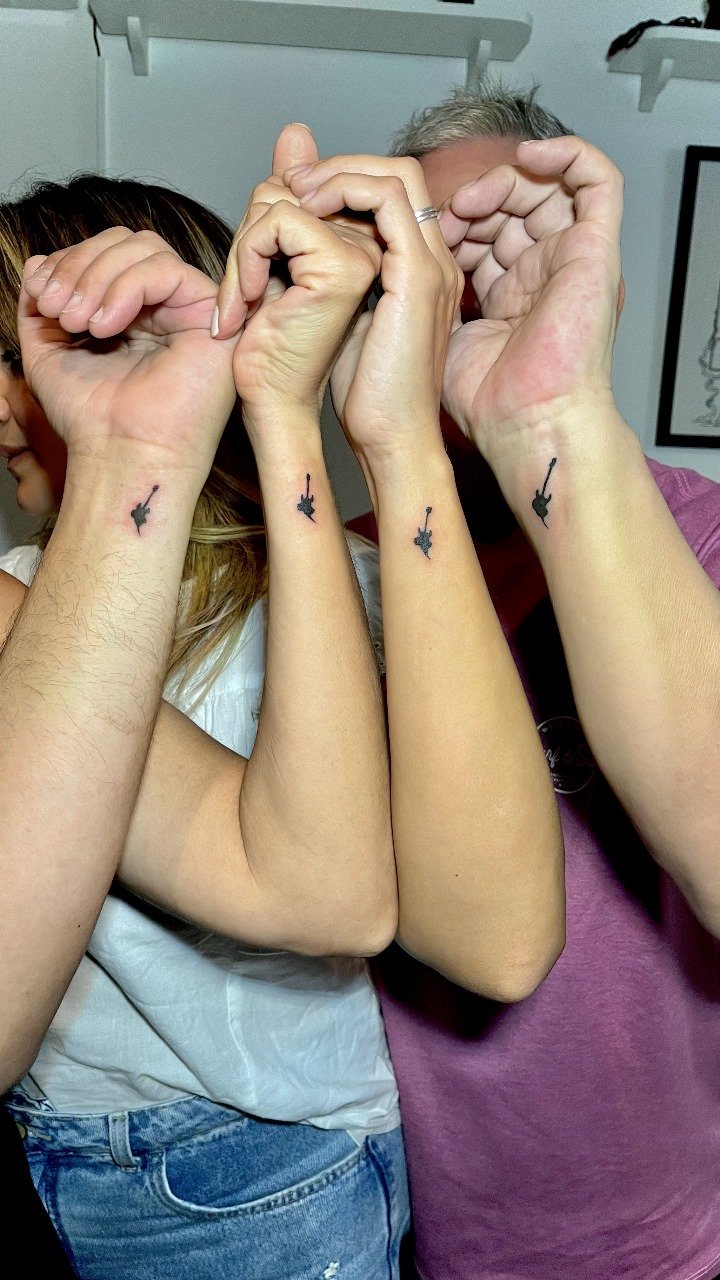 Duas pessoas com tatuagens de uma guitarra elétrica na parte interna do pulso, fazendo um coração com as mãos.