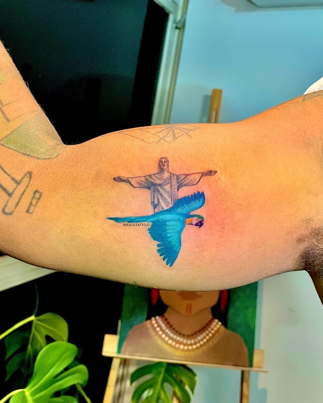 Tatuagem de uma figura que lembra Jesus com braços abertos, acompanhada de uma arara azul com bico alaranjado, na parte superior do braço de uma pessoa.