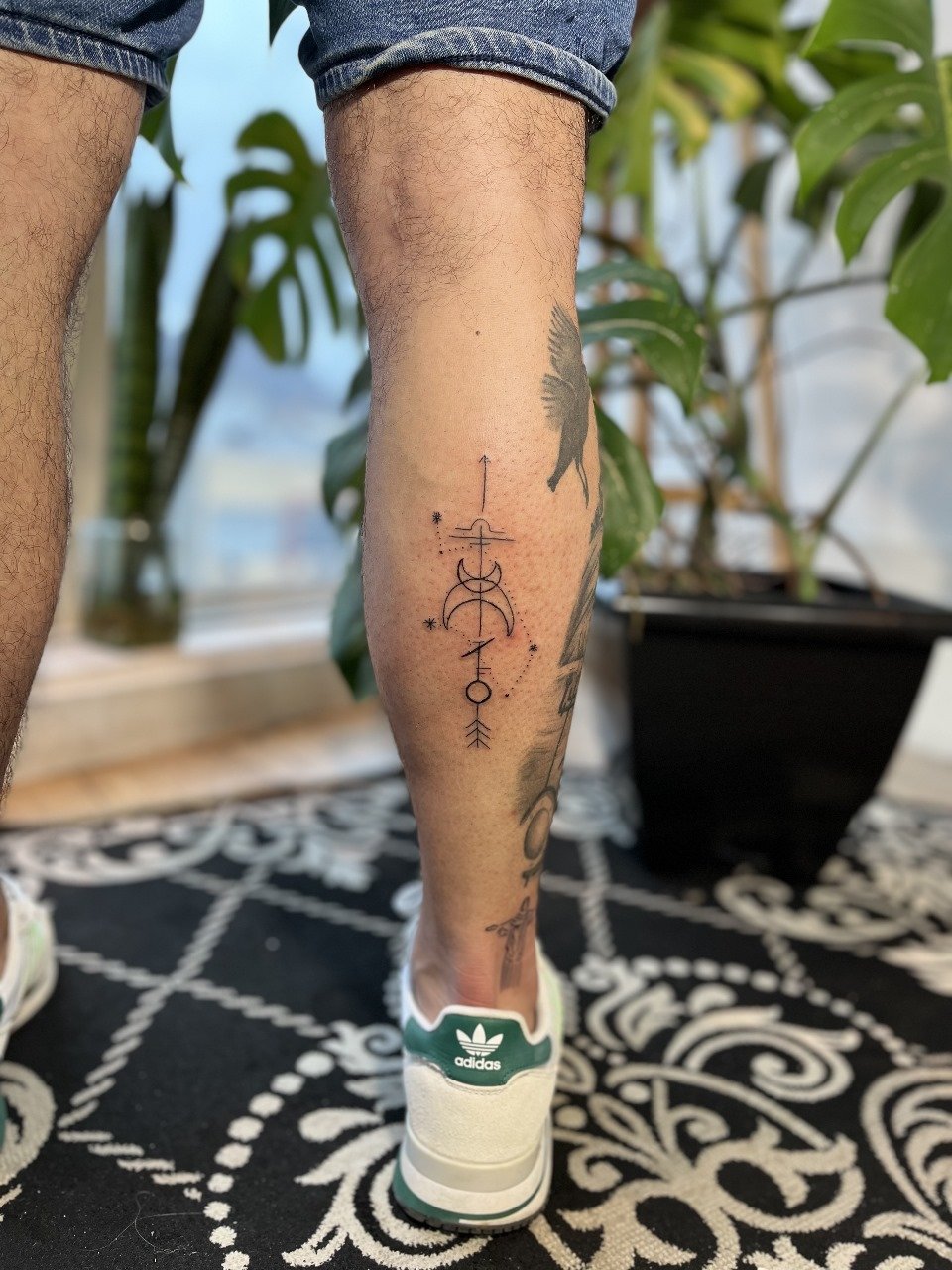 Pessoa com tatuagem na perna, vestindo shorts jeans e tênis adidas branco com detalhes verdes, em local com plantas ao fundo e tapete preto com branco.