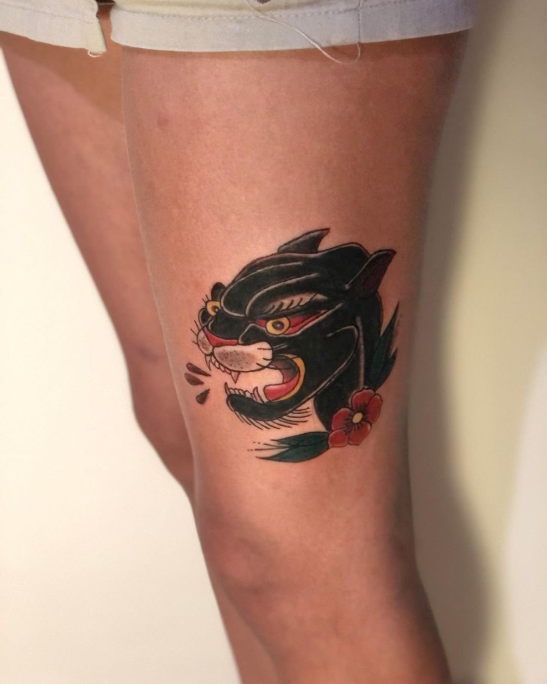 Tattoo de uma pantera preta com detalhes dourados, vermelhos e brancos, acompanhada de uma flor vermelha com folhas verdes, localizada na parte superior da coxa de uma pessoa.