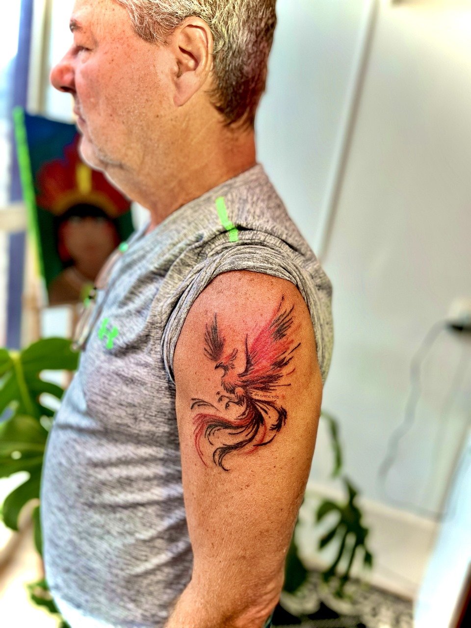 Tatuagem de um dragão estilizado em preto e vermelho no braço de um homem idoso com camisa cinza. Background com plantas verdes e ambiente interno.