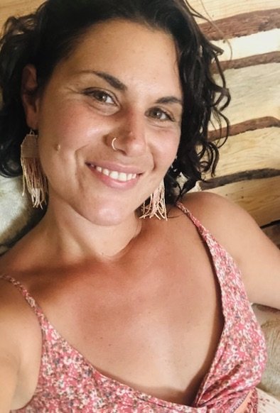 A woman with dark curly hair, wearing a pink floral top and feather earrings, smiling at the camera indoors with wooden walls in the background.