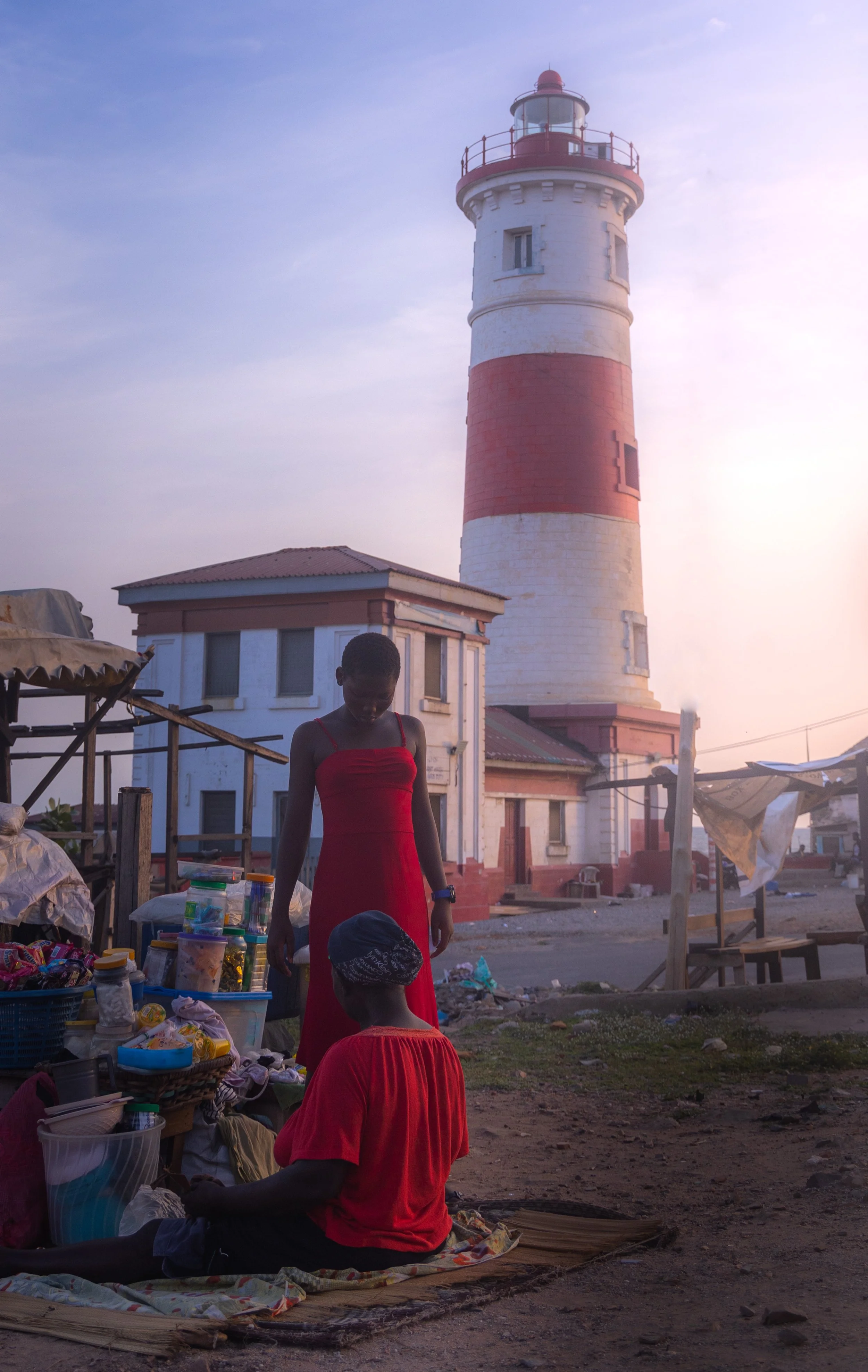 Two Ghanaian women dressed in red, under the shadow of the Jamestown lighthouse during sunset - Jamestown, Ghana.