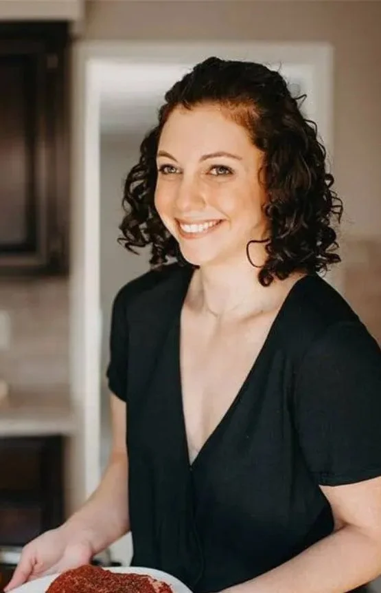 A young woman with curly brown hair and fair skin smiling while holding a plate of food in a kitchen.