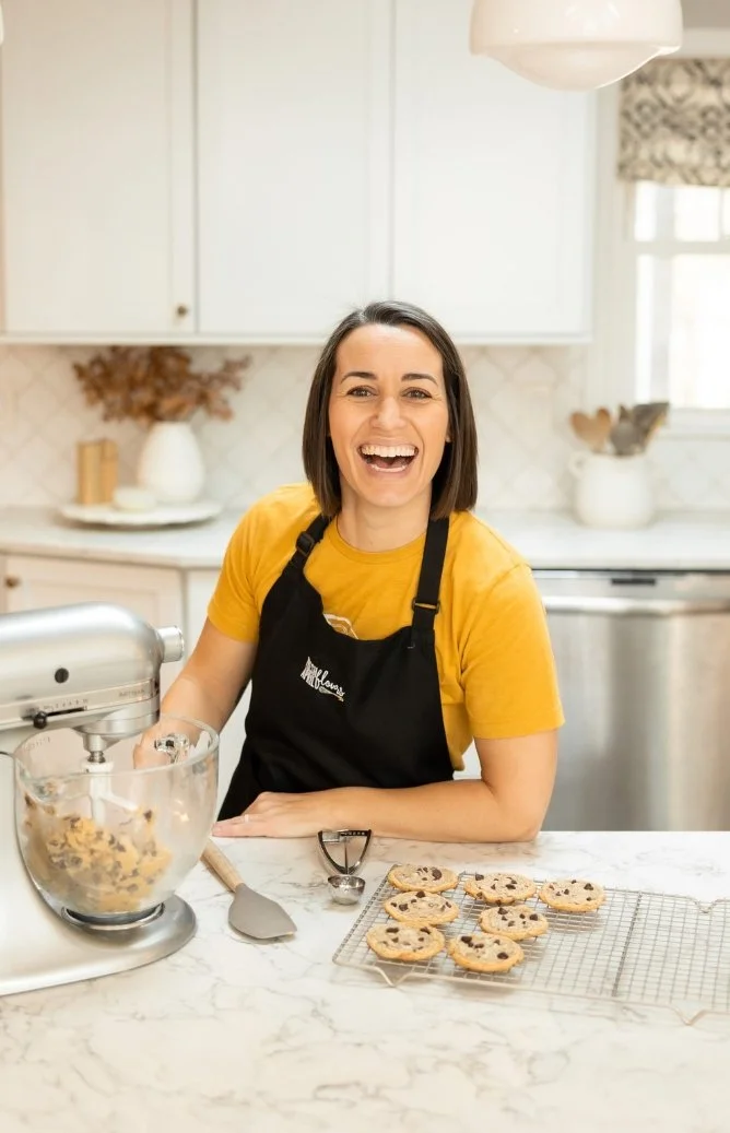 Happy woman wearing a black apron and yellow shirt, standing in a bright kitchen, with freshly baked chocolate chip cookies on a cooling rack and cookie dough in a stand mixer bowl.