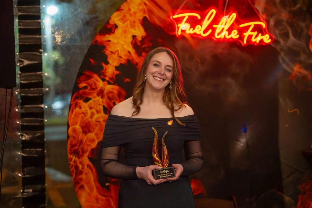 A woman with long hair in a black off-shoulder dress smiling while holding an award plaque, standing in front of a fiery backdrop with a neon sign that says 'Fuel the Fire'.