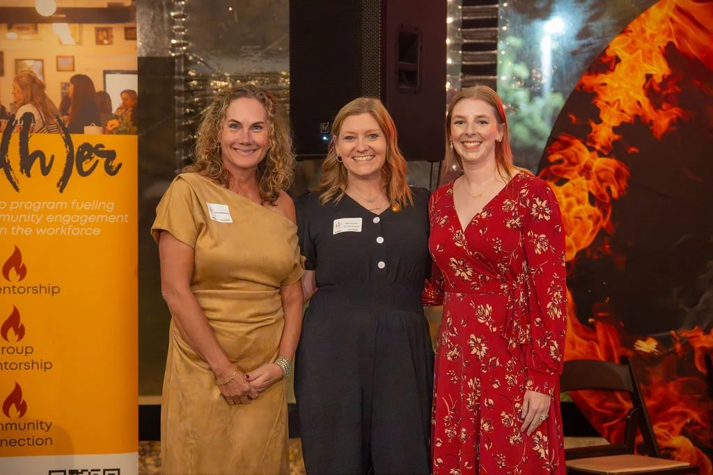 Three women smiling and posing together at an indoor event or conference, with a large fire-themed decoration and promotional banner in the background.
