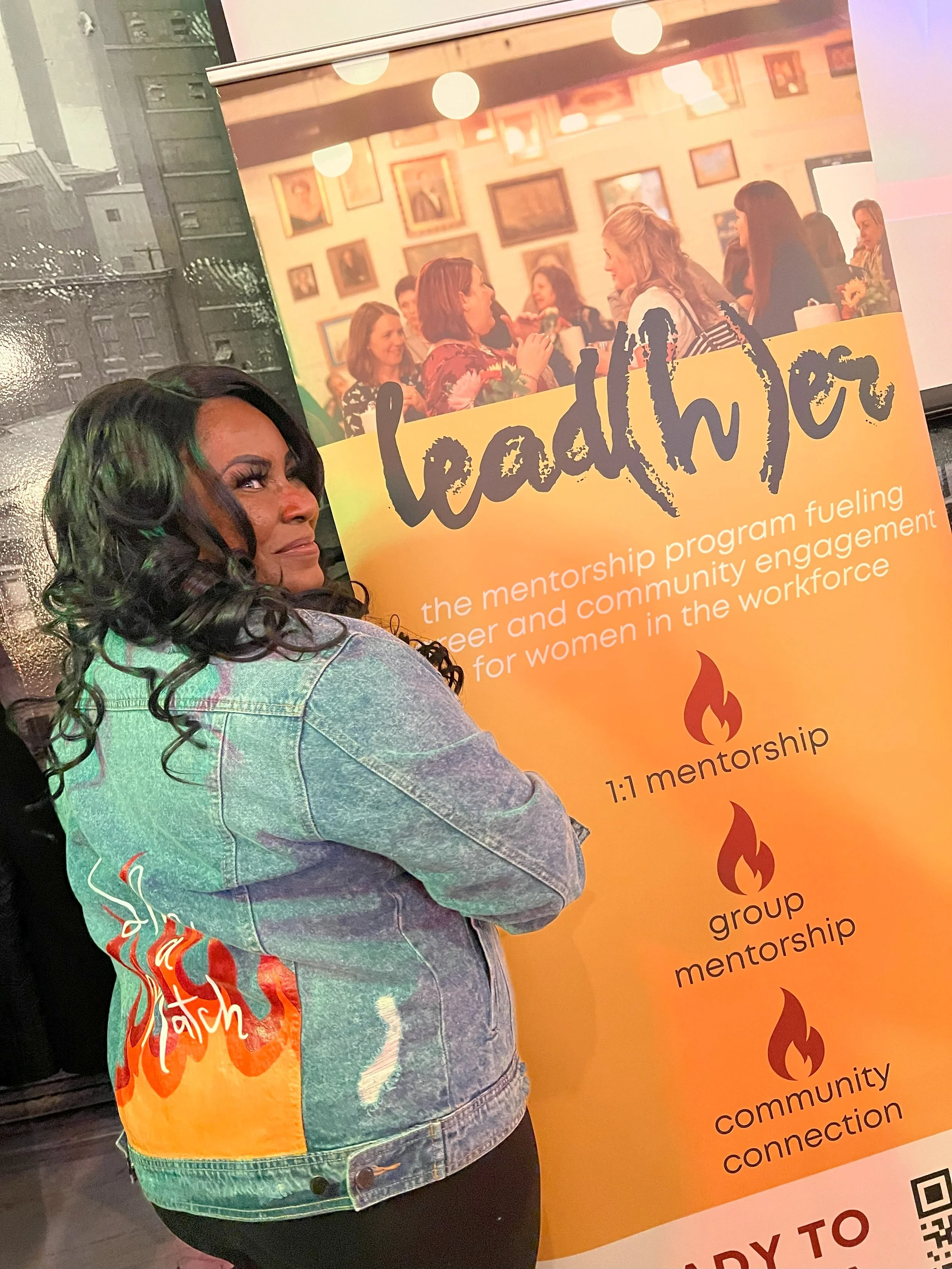 A woman with long, dark, curly hair wearing a denim jacket standing at an event next to a large orange and yellow poster. The poster displays an image of women at a social gathering and text about mentorship, community engagement, and workforce development.