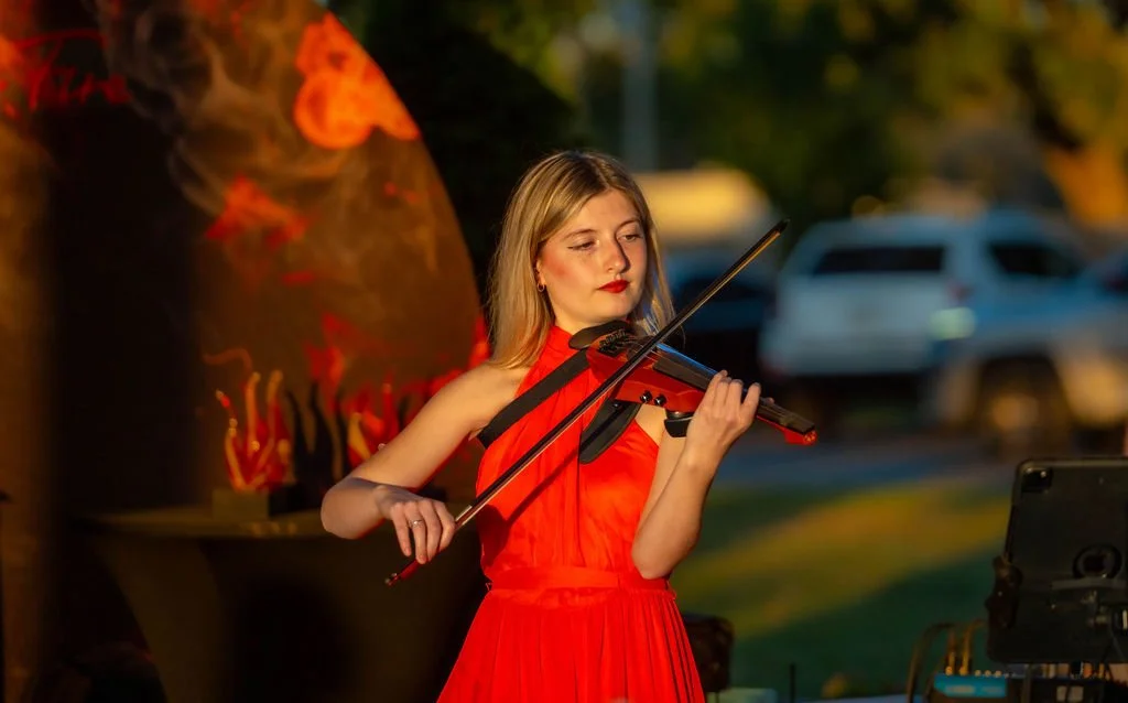 A young woman in a red dress playing the violin outdoors at sunset.