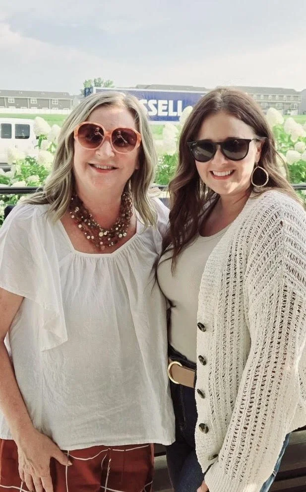 Two women standing together outdoors, smiling. One woman has blonde hair, wearing a white blouse, coral sunglasses, and layered necklaces. The other woman has brown hair, wearing black sunglasses, a white shirt, and a cream-colored knitted cardigan. They are in front of a lush green background with large white flowers, and a