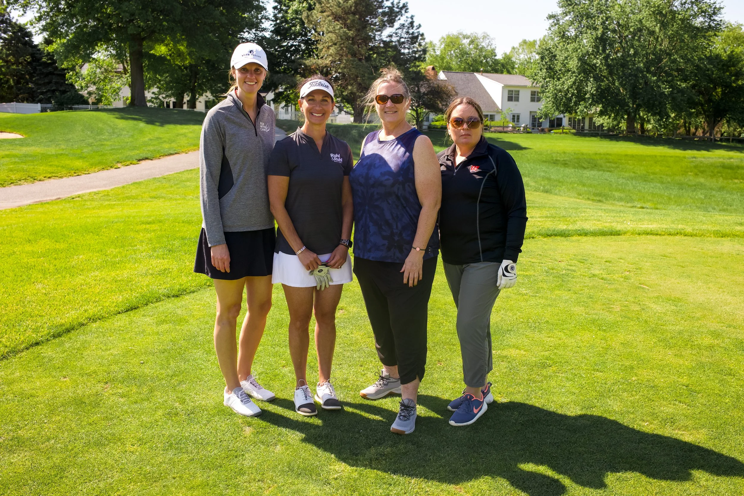 Four women standing on a golf course, smiling and posing for the photo, with trees and houses in the background.