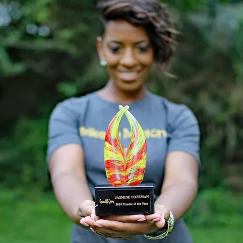 Woman holding a colorful trophy with a black plaque that reads 'Jasmine Bozeman, 2018 Mentor of the Year' in an outdoor setting.