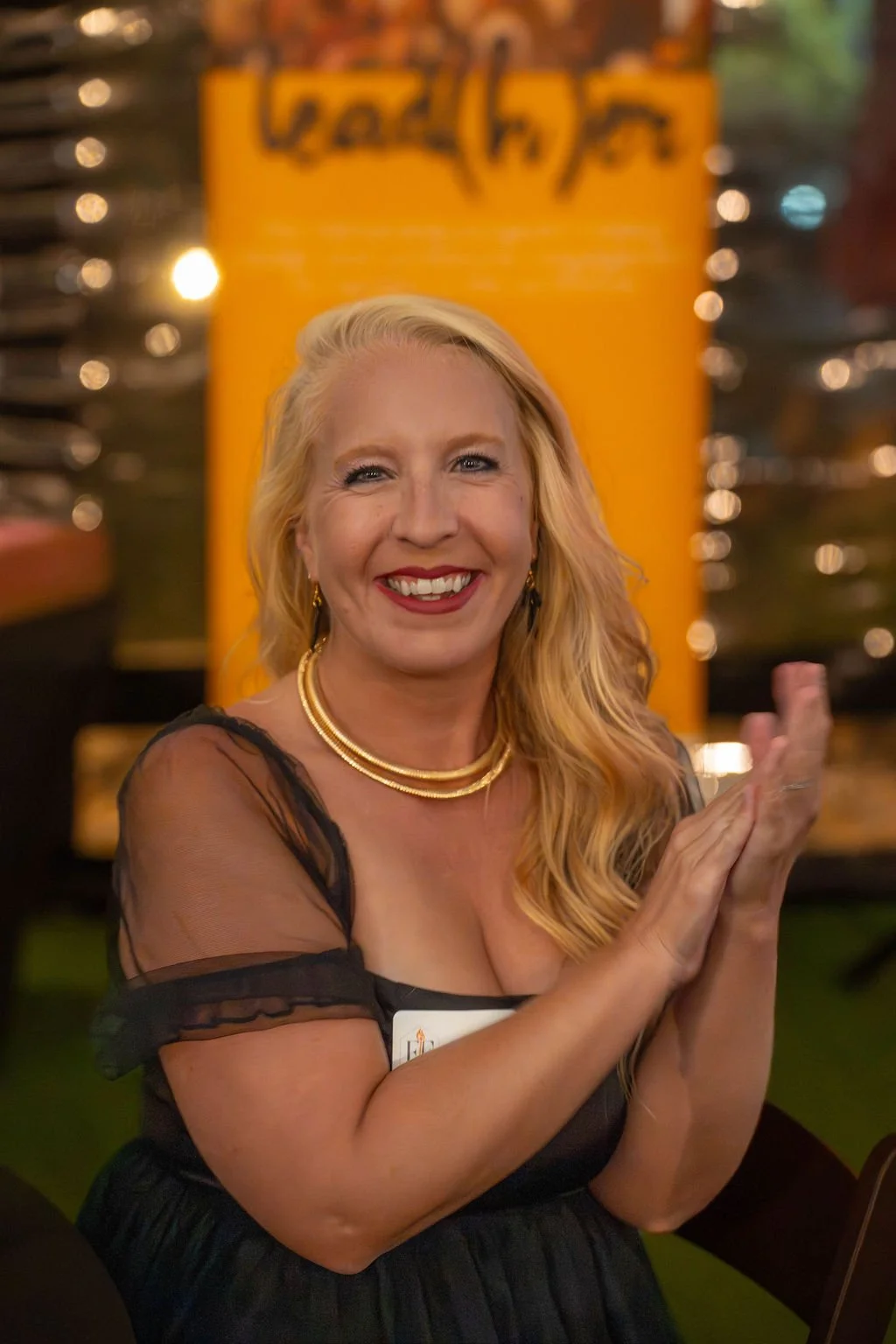 A smiling woman with blonde hair, wearing a black dress with sheer sleeves, gold jewelry, and clapping her hands, standing in front of a yellow background with a black logo.