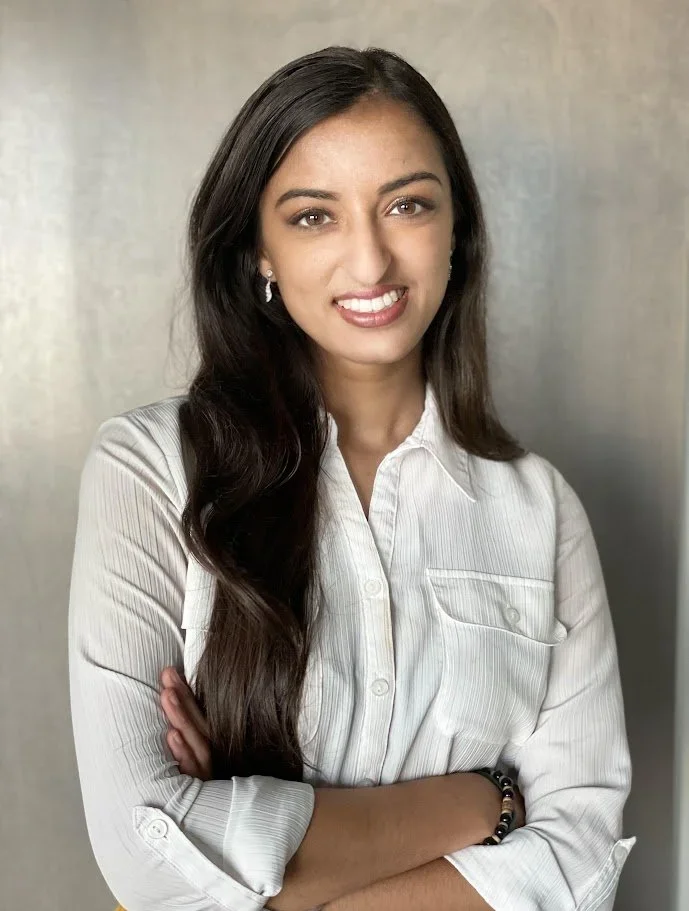 A young woman with long dark hair, wearing a white button-up shirt, standing with arms crossed and smiling at the camera.