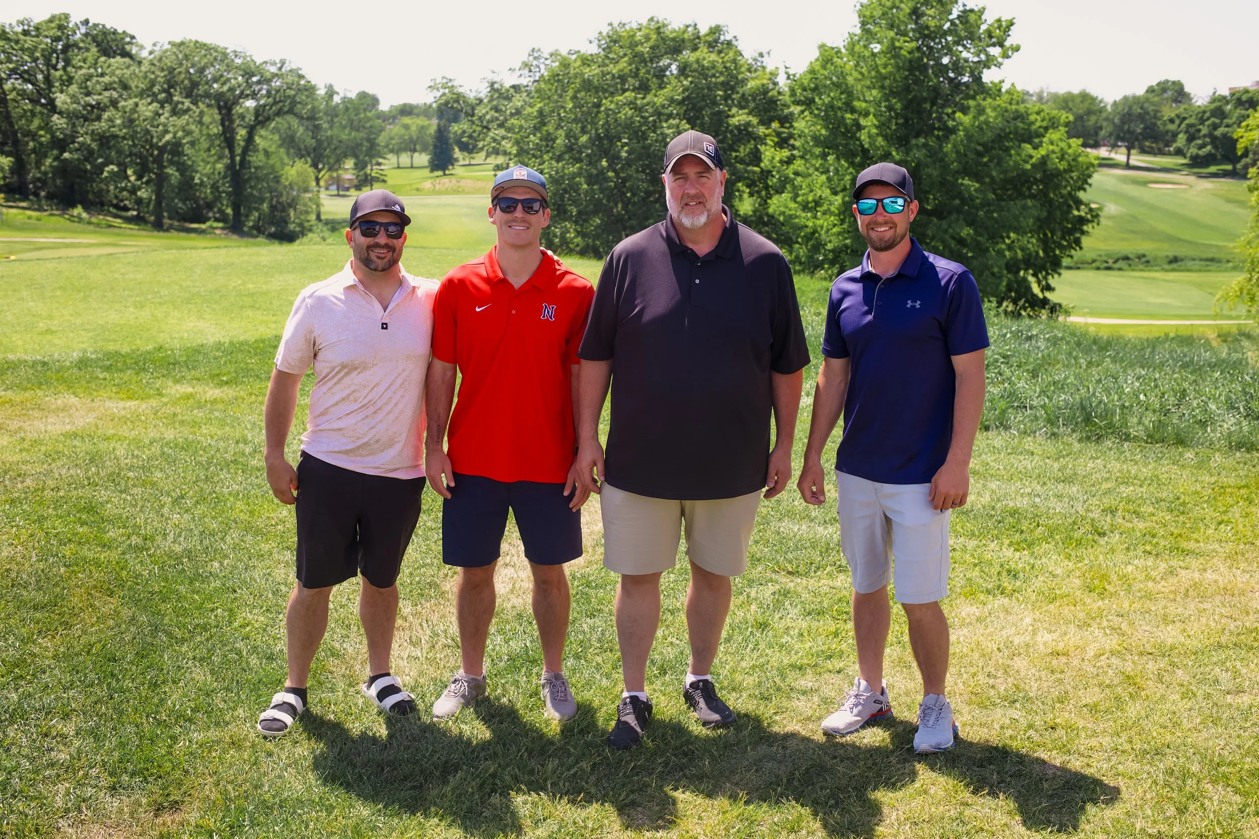 Four men standing outdoors on a sunny day on a grassy area with trees and a golf course in the background. They are dressed casually, some wearing sunglasses and baseball caps.