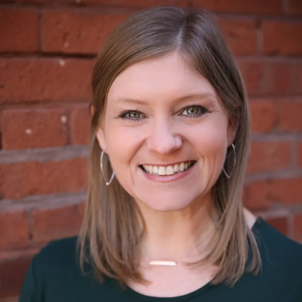 Close-up portrait of a smiling woman with shoulder-length brown hair wearing hoop earrings and a black top, standing against a brick wall.